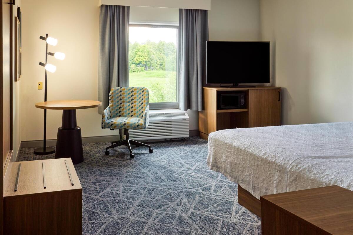A hotel room with a bed, a desk with a lamp, a chair, a TV, and a view of the outdoors.