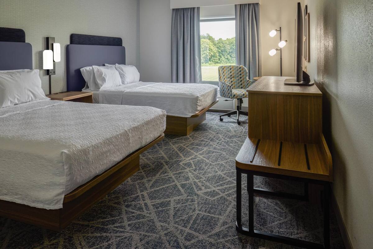 A hotel room with two beds, a desk, a chair, and a large window with curtains.