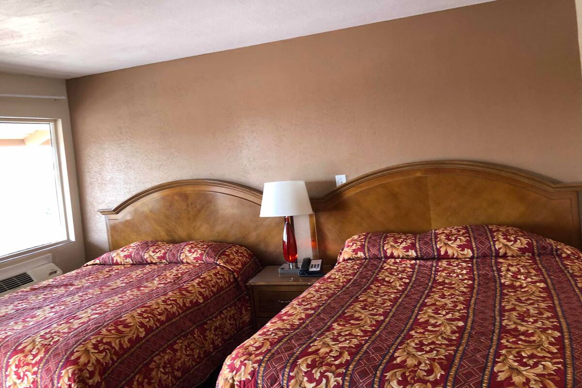 A hotel room with two beds, a nightstand with a lamp, and a window providing natural light.