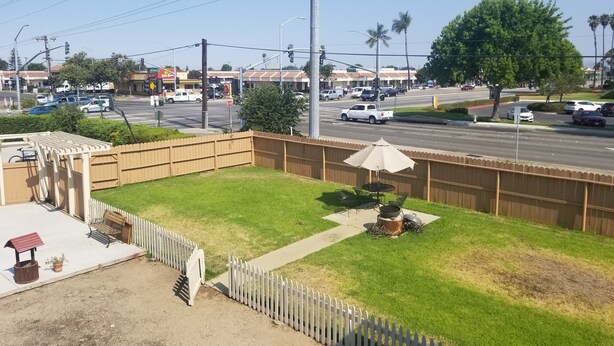 Ein eingezäunter Garten mit Terrasse, einem kleinen Gemüsegarten und Blick auf eine belebte Straße mit Autos und Palmen.