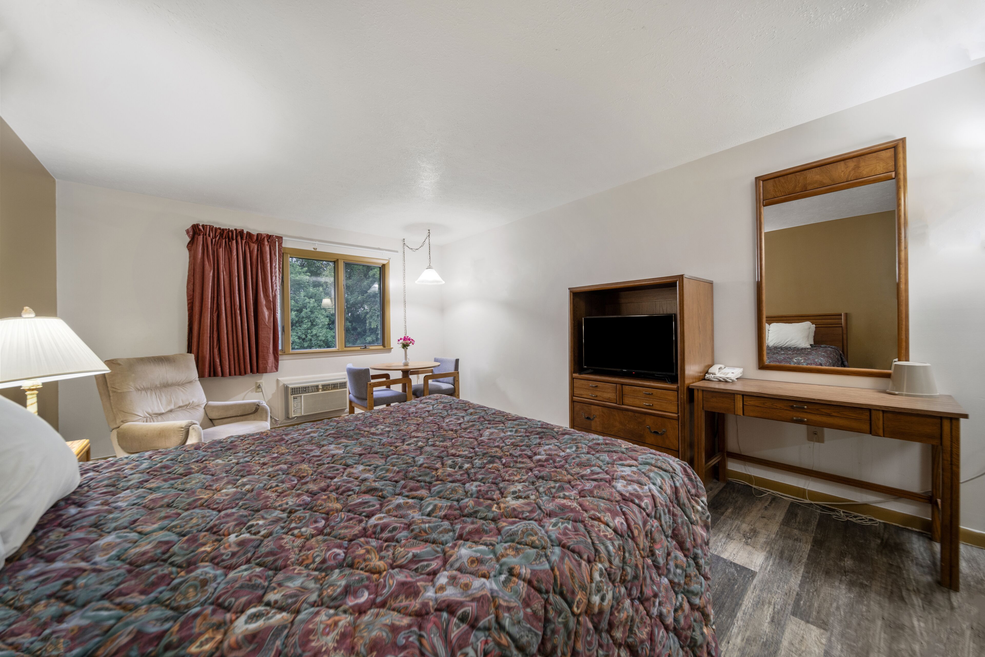 A hotel room with a bed, a desk, a chair, a TV, a mirror, and a window with curtains.