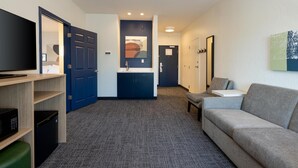 Habitación de hotel con televisor de pantalla plana, kitchenette, sofá gris y una pequeña mesa.