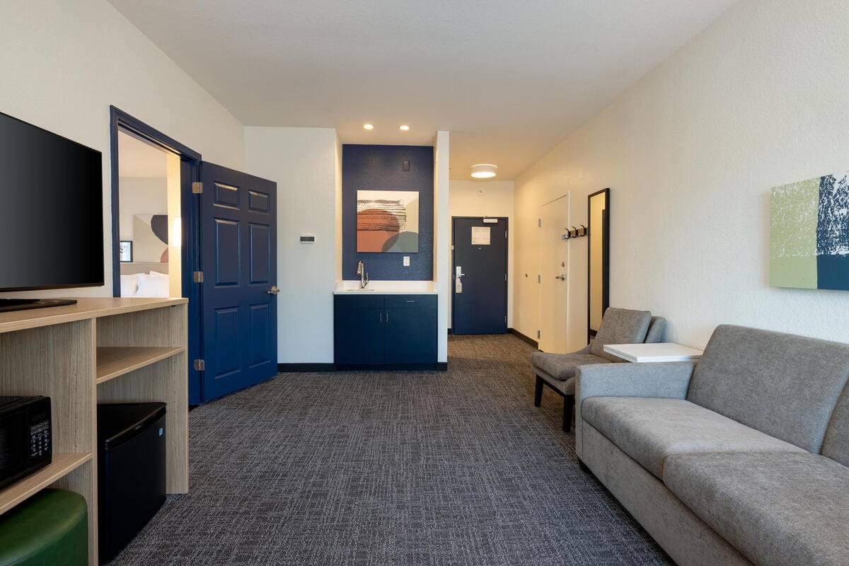 A hotel room with a flat-screen TV, a kitchenette, a grey sofa, and a small table.