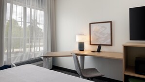 Habitación de hotel con cama, escritorio con lámpara, silla, un cuadro enmarcado y televisión sobre una estantería.