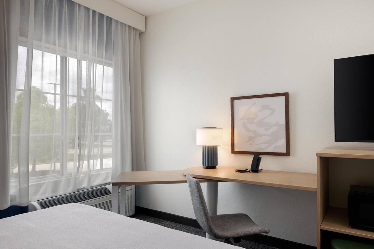 A hotel room with a bed, a desk with a lamp, a chair, a framed picture, and a TV on a shelf.