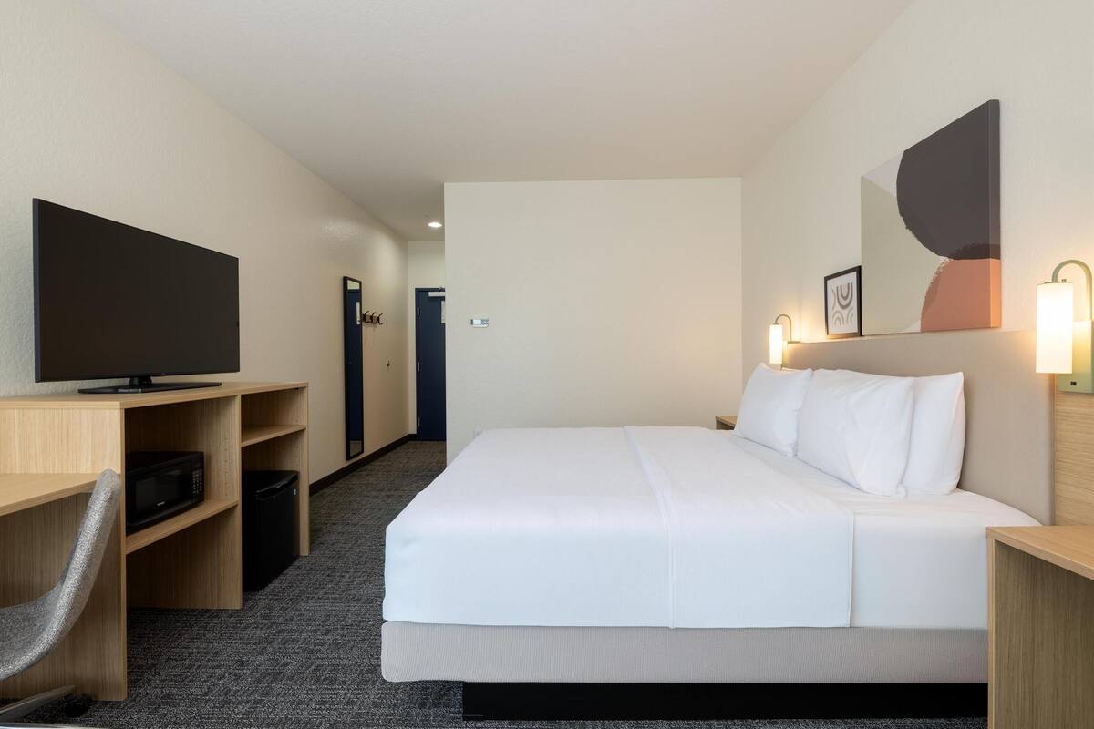 A hotel room with a bed, a TV on a wooden shelf, a microwave, and a trash bin.