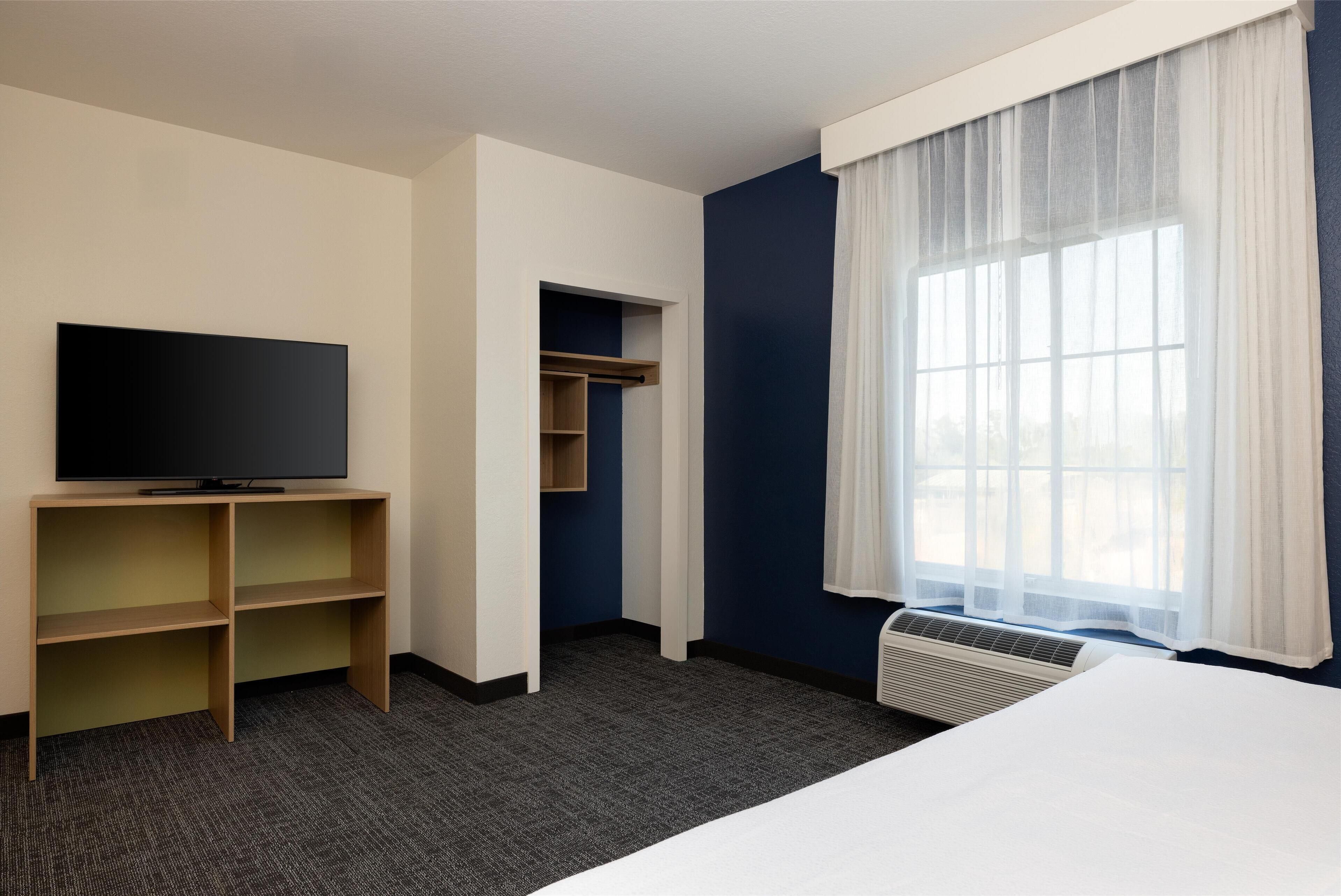 A hotel room with a flat-screen TV, built-in shelving, a bed, a window with sheer curtains, and a wall-mounted air conditioning unit.