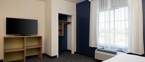 A hotel room with a flat-screen TV, built-in shelving, a bed, a window with sheer curtains, and a wall-mounted air conditioning unit.