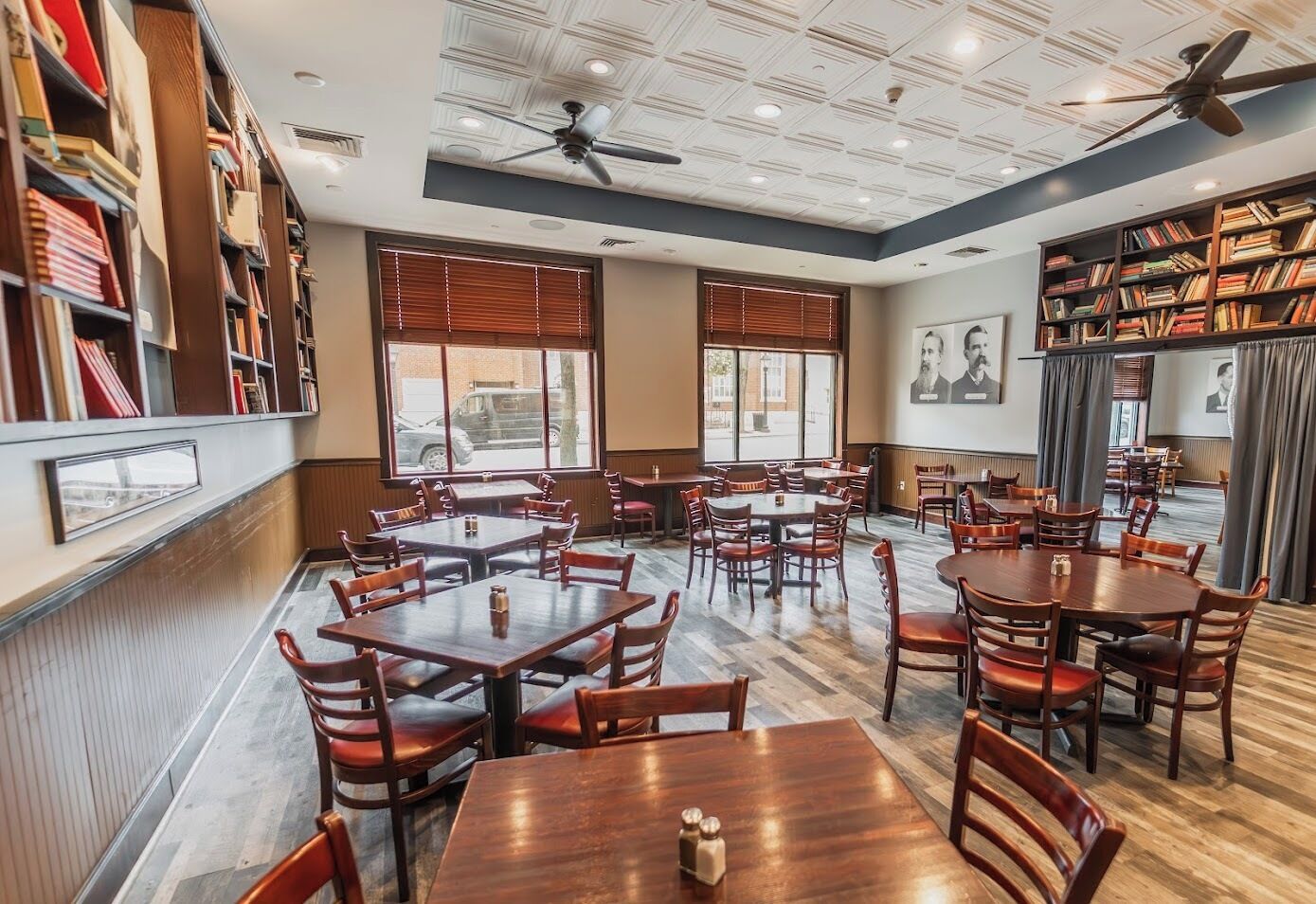 Un ristorante con diversi tavoli e sedie, ampie finestre, ventilatori a soffitto e scaffali di libri.