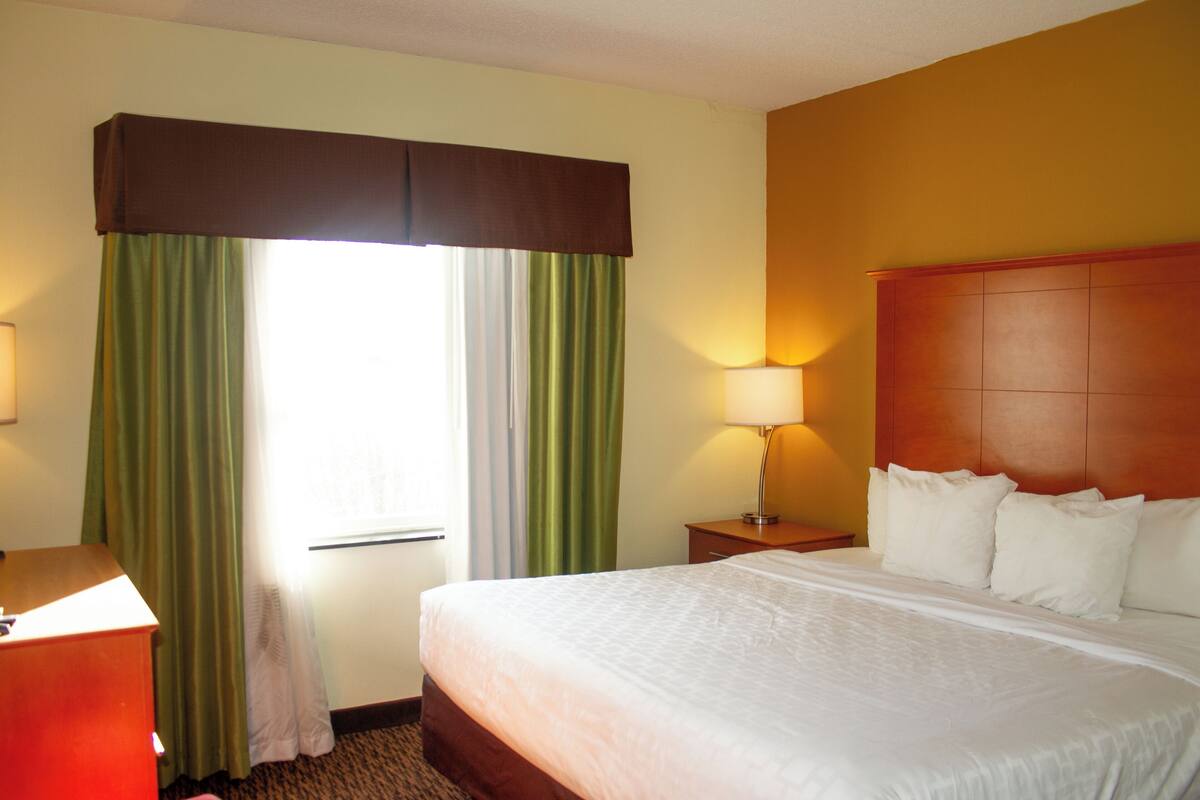 A hotel room with a bed, a nightstand, a lamp, a window with curtains, and a wall with a headboard.