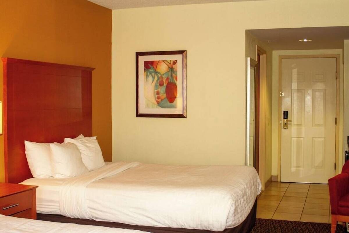 A hotel room with a bed, a nightstand, a framed picture on the wall, and a door leading to another room.
