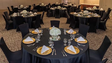 A banquet hall with round tables set for dining, each with gold plates, white napkins, and floral centerpieces.