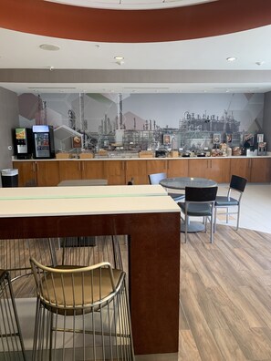 Un café moderne avec un comptoir central, un espace salon et une fresque murale représentant un paysage urbain industriel.