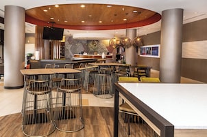 A modern restaurant with high tables and metal chairs, a bar area, and a large TV screen.