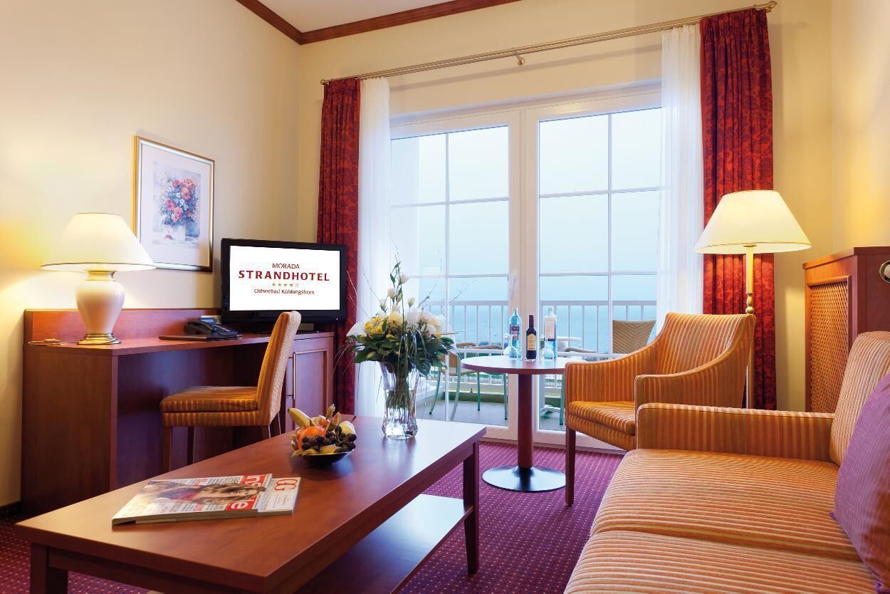 A hotel room with a television, a desk with a magazine, a sofa, and a balcony with a view.