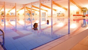 Uma grande piscina interior com água cristalina, rodeada por espreguiçadeiras e camas de banho, e um teto alto com vigas de madeira.