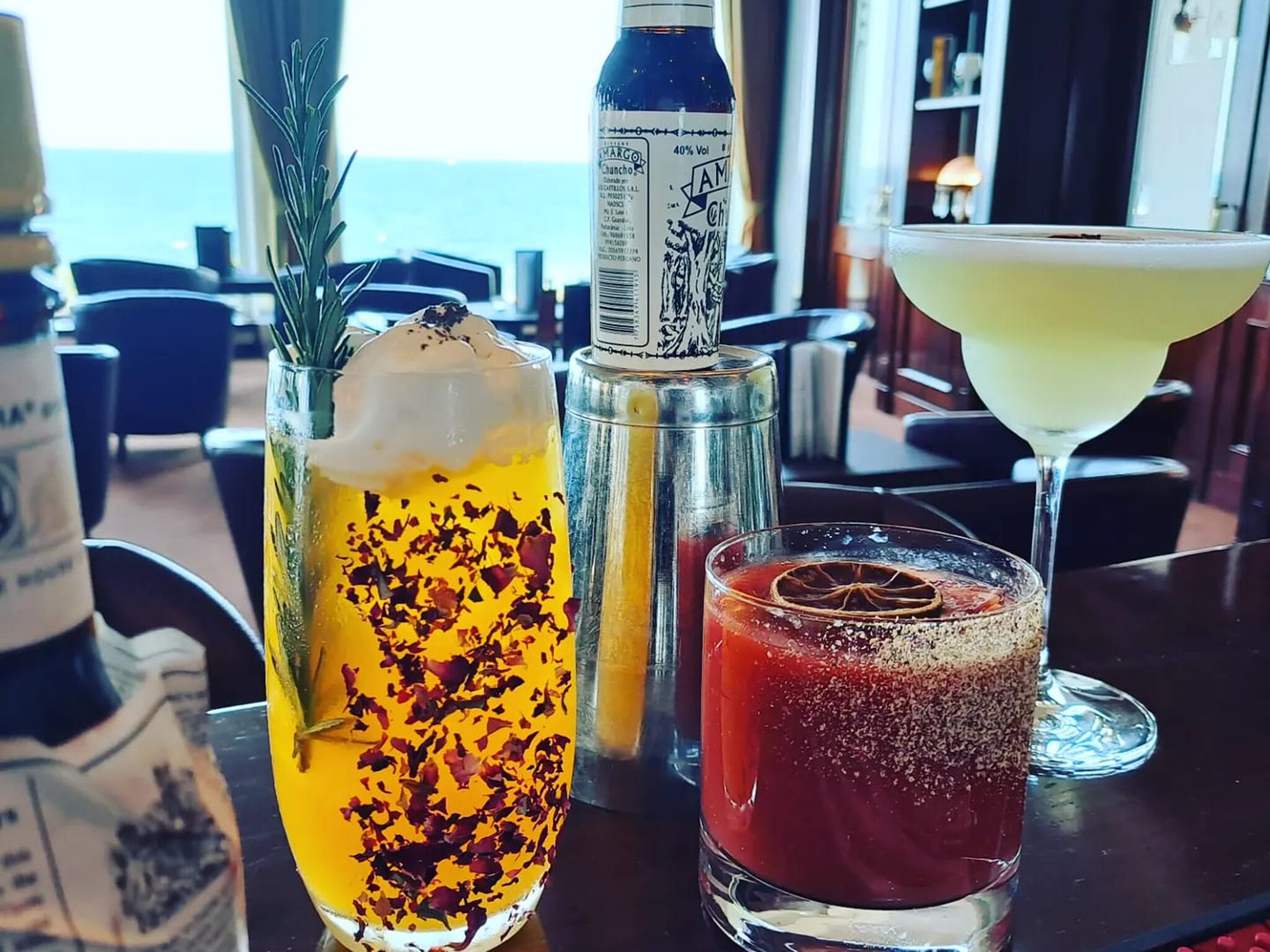 A table with three drinks: a yellow cocktail with rose petals, a red cocktail with a dried orange slice, and a clear cocktail in a martini glass.