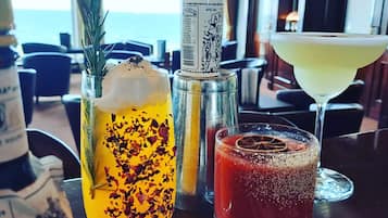 A table with three drinks: a yellow cocktail with rose petals, a red cocktail with a dried orange slice, and a clear cocktail in a martini glass.