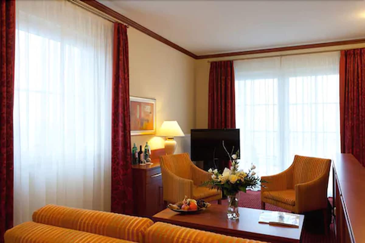 Ein Hotelzimmer mit einer Couch, zwei Sesseln, einem Couchtisch mit Blumen, einem Fernseher und einer Lampe.