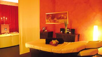 A hotel room with a bed, a red curtain, a painting, a vase with flowers, a bedside table with a lamp, and a small table with a speaker and a candle.