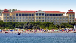 Um hotel grande com várias varandas e telhado vermelho, situado perto de uma praia com pessoas nadando e tomando sol.