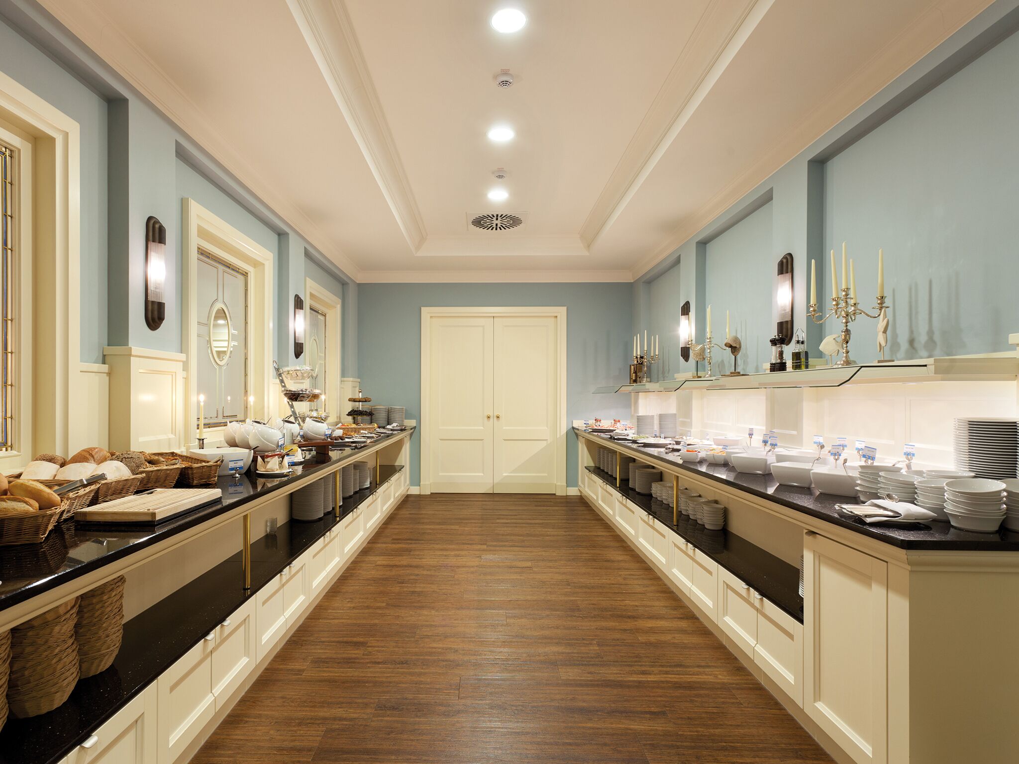 A long, narrow hotel room with a central door, two side counters with breakfast items, and a chandelier.