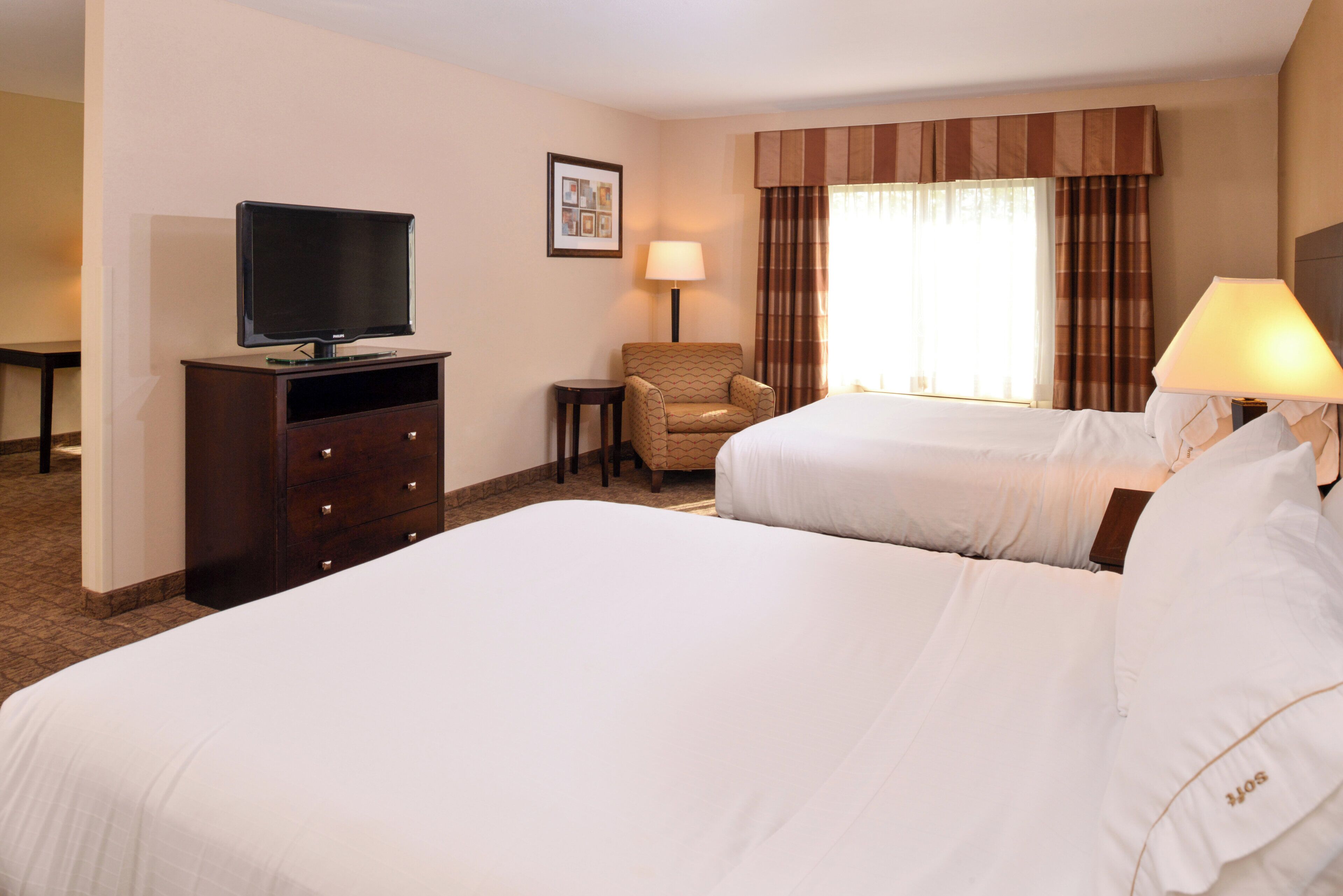 Une chambre d’hôtel avec deux lits, une télévision, une chaise, une lampe et une fenêtre avec des rideaux.