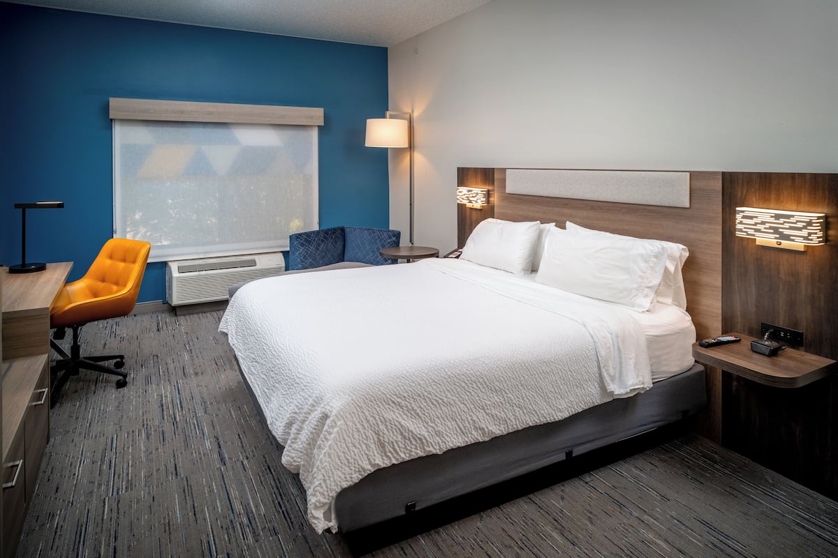 A hotel room with a large bed, a desk with a chair, a window with blinds, a standing lamp, and a wall-mounted light fixture.