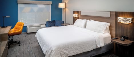 Hotellihuone, jossa on suuri sänky, työpöytä tuolilla, ikkuna, jossa on sälekaihtimet, lattiavalaisin ja seinään kiinnitetty valaisin.