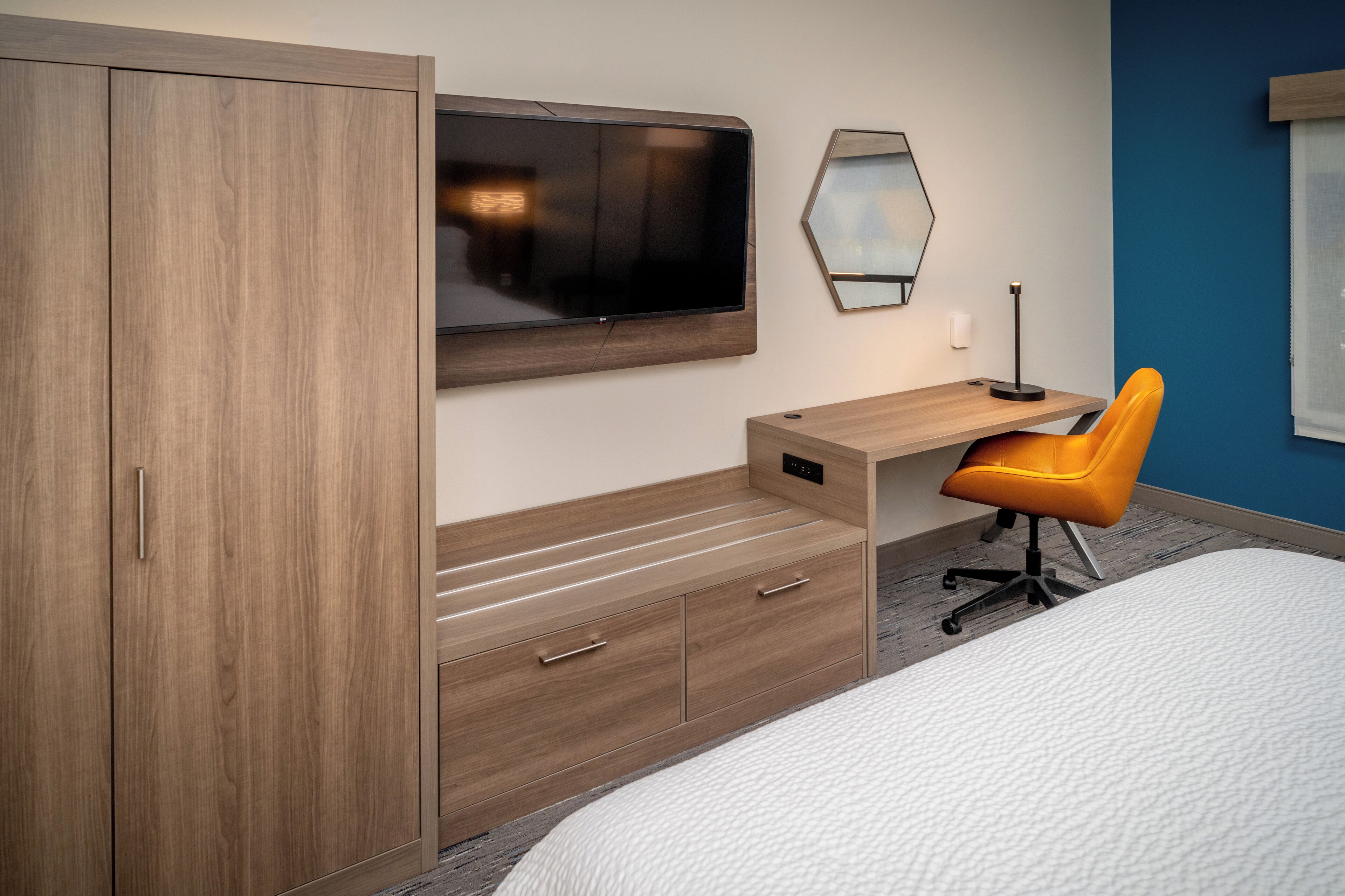 A hotel room with a wooden wardrobe, a desk with a chair, a flat-screen TV, and a mirror on the wall.