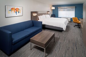 Quarto de hotel moderno com sofá azul, mesa de centro de madeira, cama com roupa de cama branca, escrivaninha com cadeira e uma janela grande com persianas.