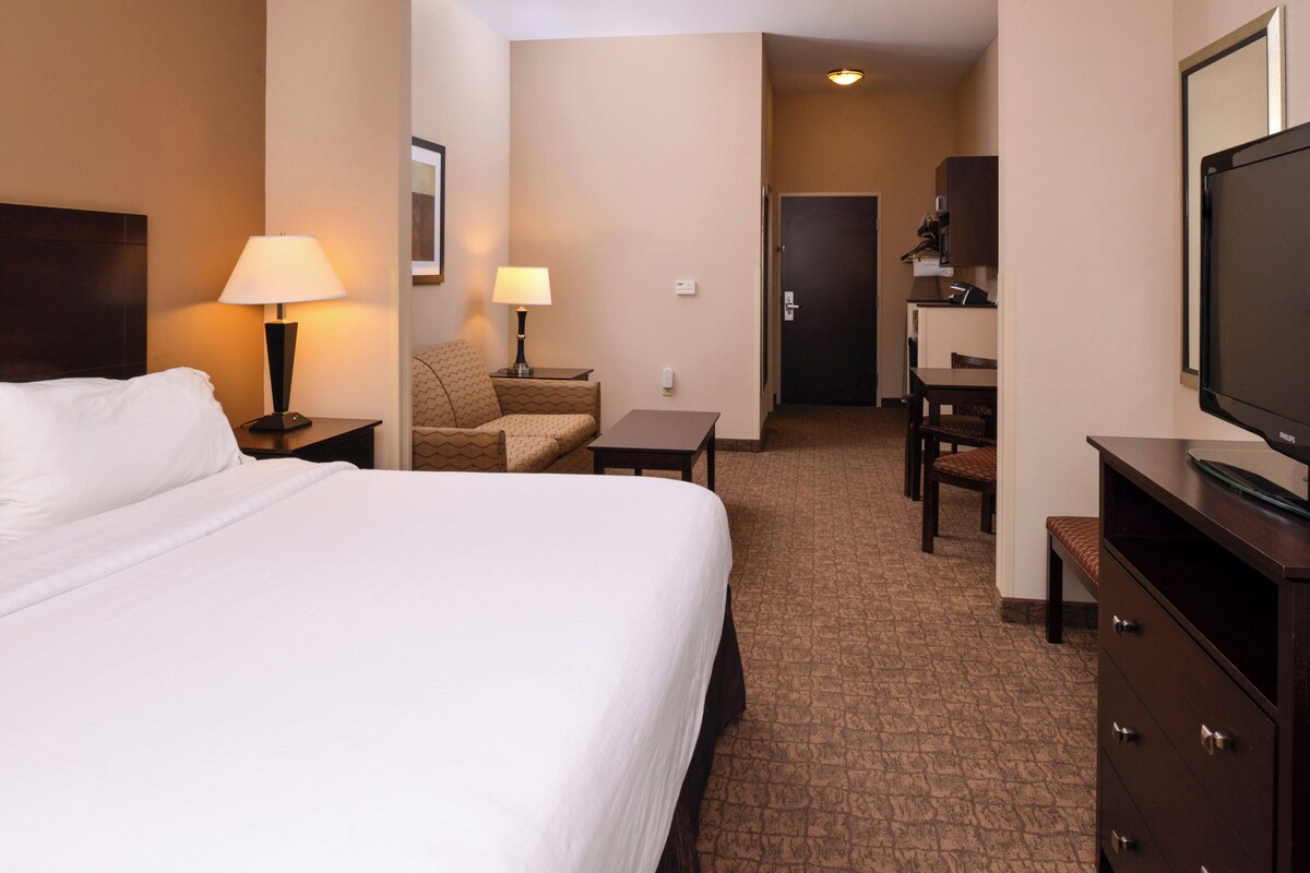 Habitación de hotel con cama, escritorio, silla, lámpara, televisor y puerta que da a un pasillo.