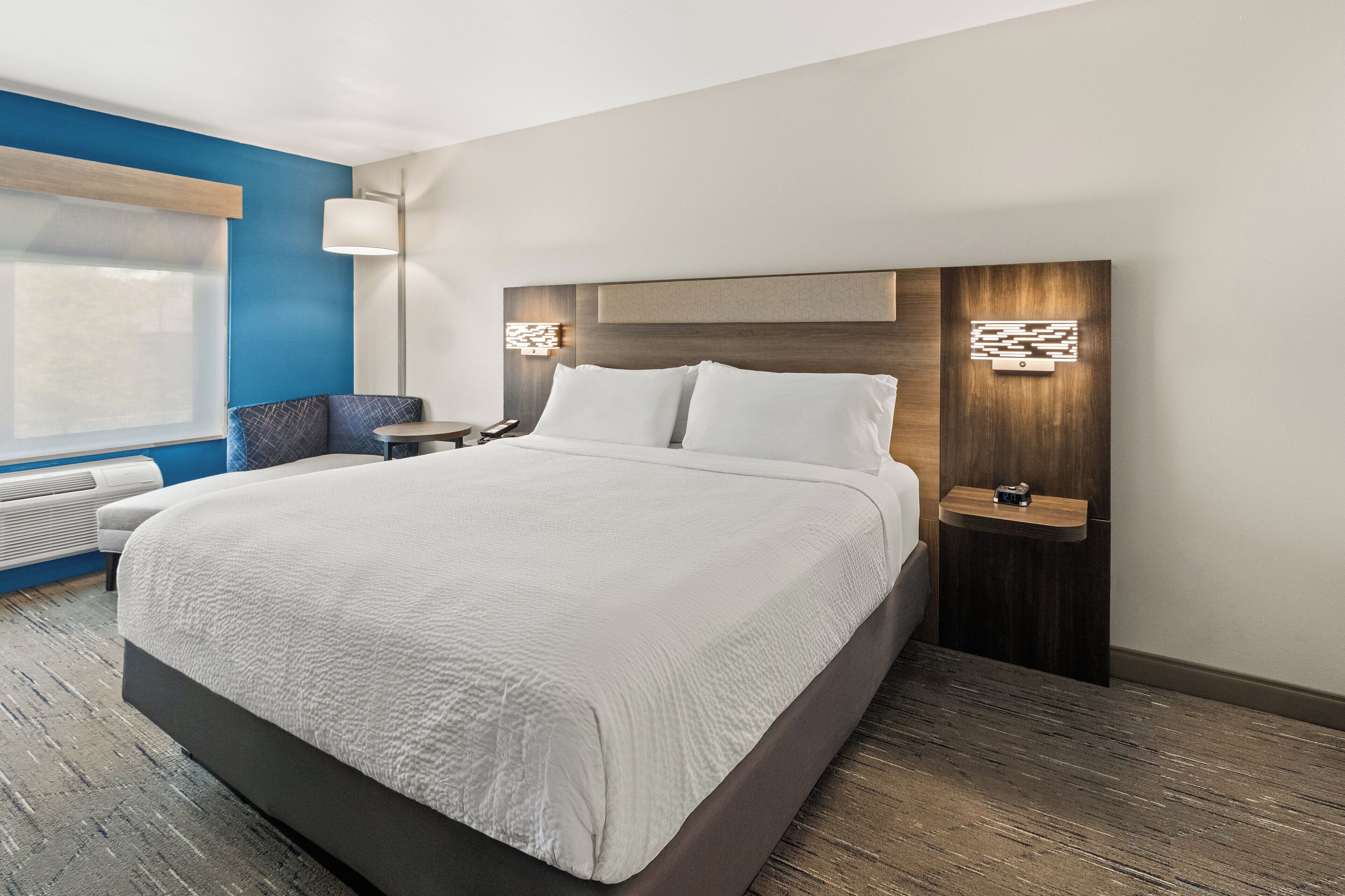 A modern hotel room with a large bed, a bedside table, a lamp, and a view of the outdoors.