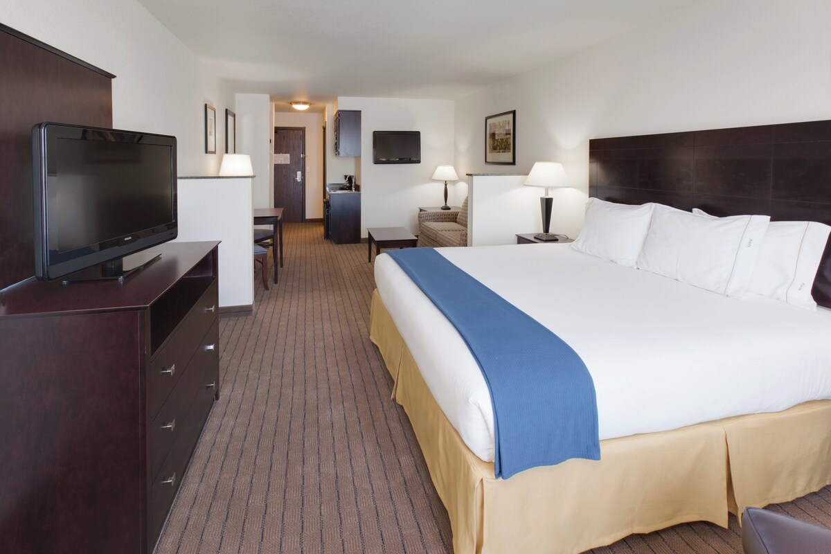 Ein Hotelzimmer mit einem großen Bett, einem Fernseher auf einer Kommode, einem Schreibtisch mit Stuhl und einer kleinen Küche im Hintergrund.
