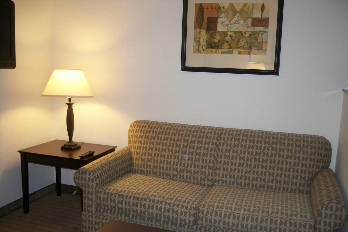 Ein Wohnzimmer mit einem gemusterten Sofa, einem kleinen Tisch mit einer Lampe und einem gerahmten Kunstwerk an der Wand.