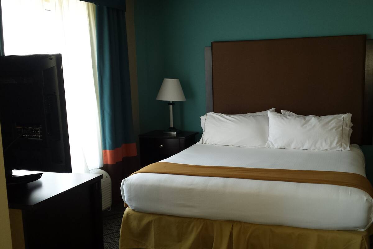 A hotel room with a large bed, two bedside tables with lamps, a TV on a stand, and a window with curtains.