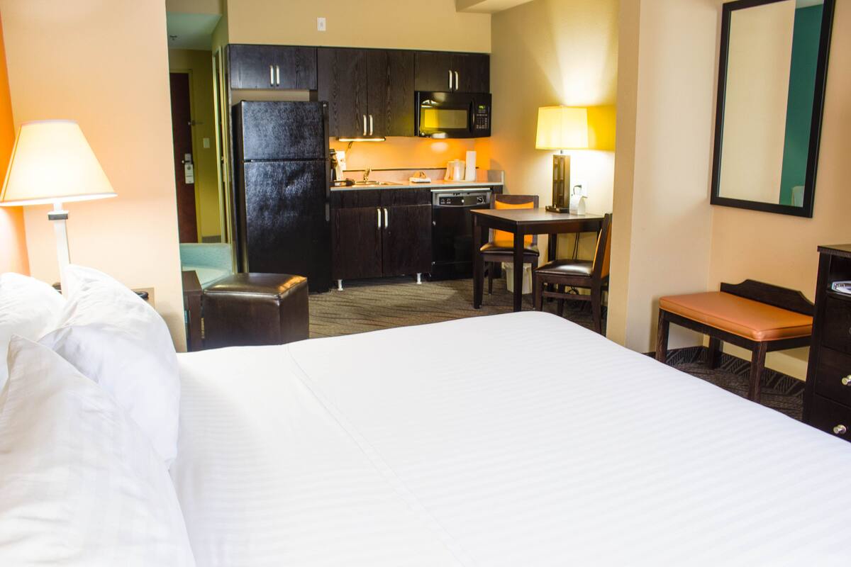 A hotel room with a bed, a desk, a chair, a small table, a bench, a microwave, and a refrigerator.