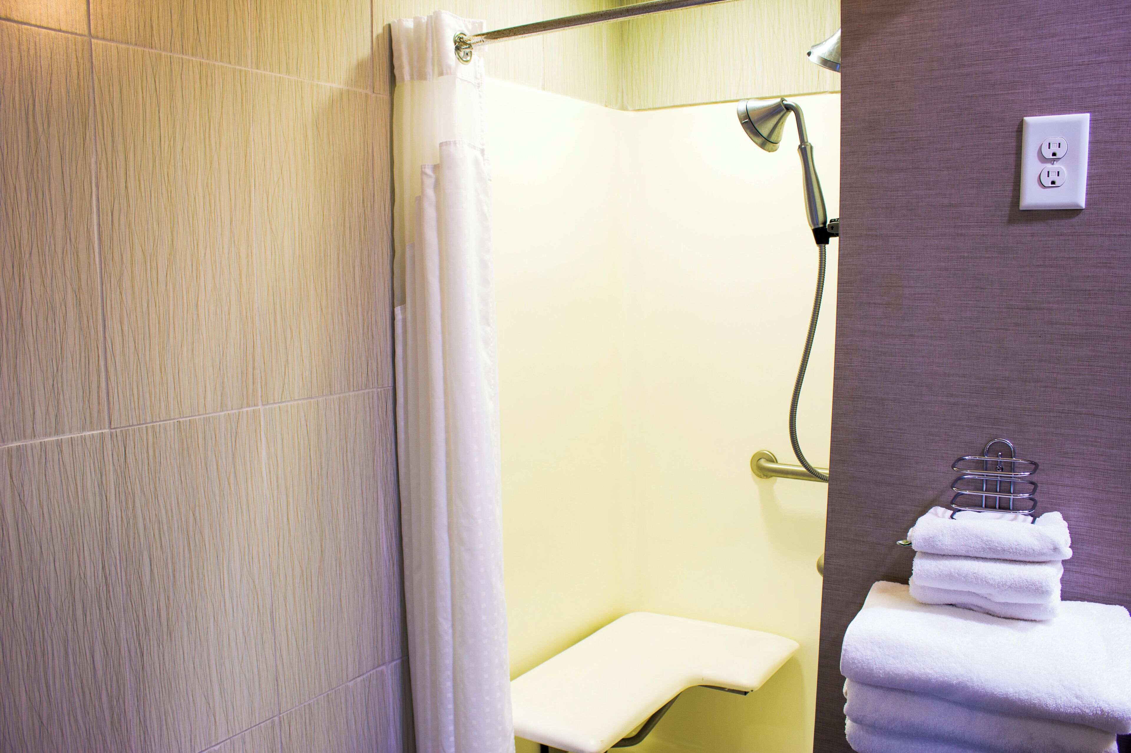 A bathroom with a shower, a towel rack with folded towels, and a power outlet on the wall.