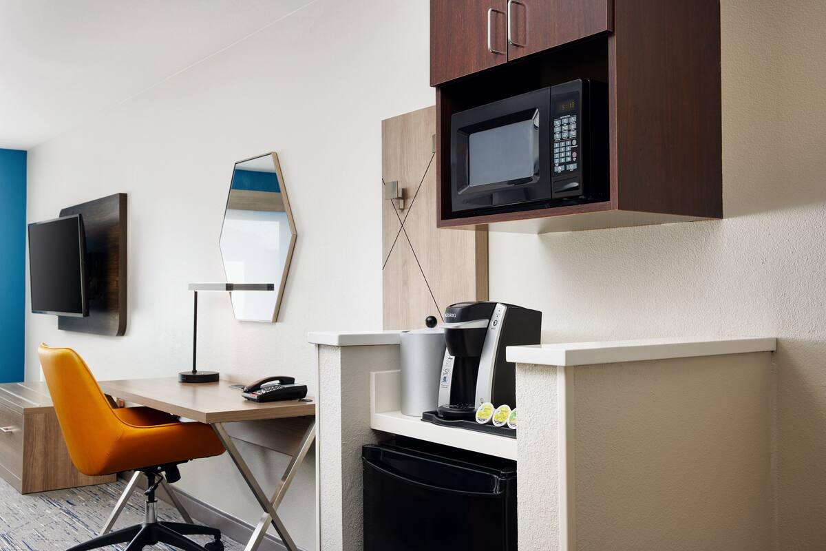Une chambre d’hôtel compacte équipée d’une kitchenette, d’un four à micro-ondes, d’une machine à café, d’une petite poubelle, d’un bureau avec une chaise et d’un téléviseur à écran plat fixé au mur.