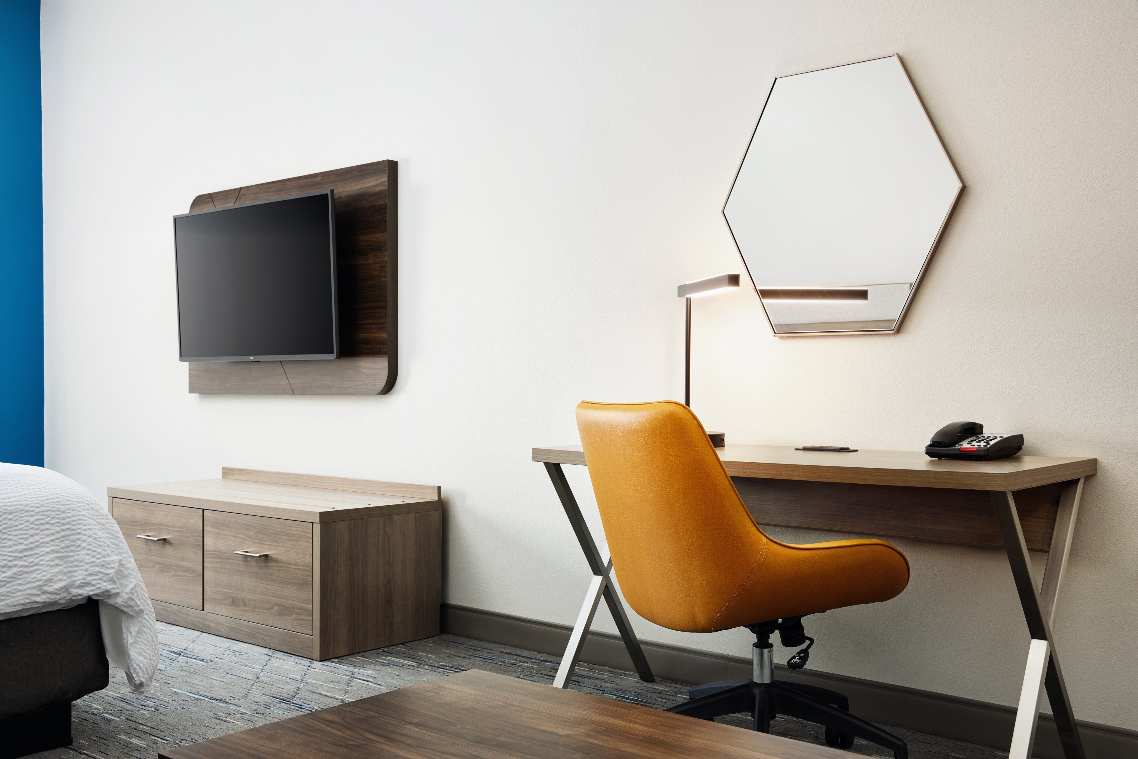 A hotel room with a bed, a wooden dresser, a desk with a chair, a flat-screen TV, and a mirror.