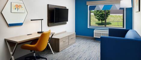 A modern room with a wooden desk, an orange office chair, a blue sofa, a flat-screen TV mounted on the wall, and a window with a view of greenery.