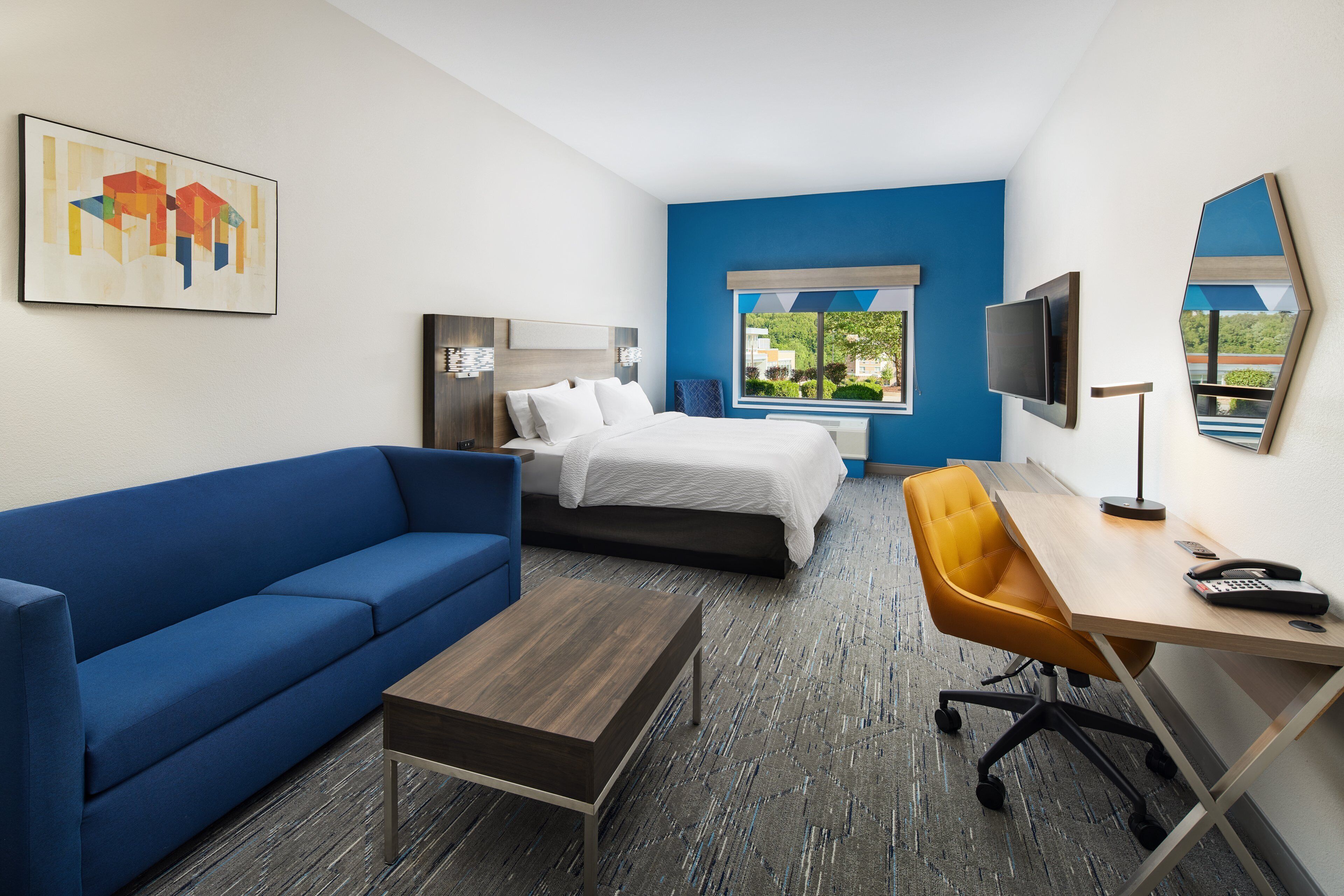 A hotel room with a blue sofa, a bed, a desk with a chair, a TV, and a window with a view of greenery.