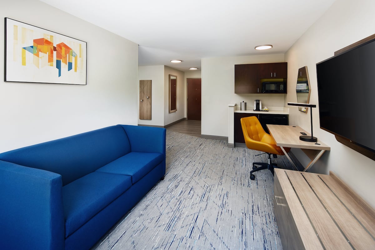 Une chambre d’hôtel moderne avec un canapé bleu, un bureau en bois, une grande télévision et un coin cuisine.