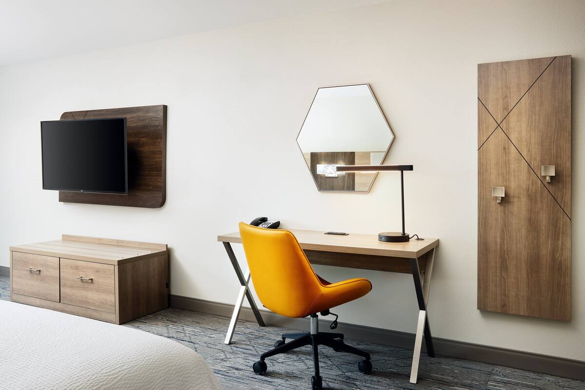 Une chambre d’hôtel avec un bureau en bois, une chaise de bureau jaune, un téléviseur à écran plat fixé au mur, une table de chevet et une armoire.