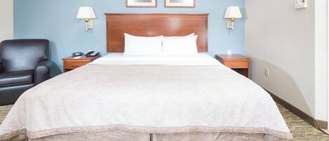 Une chambre d’hôtel avec un grand lit, une chaise, une table de chevet, deux lampes fixées au mur et deux tableaux encadrés accrochés au mur.