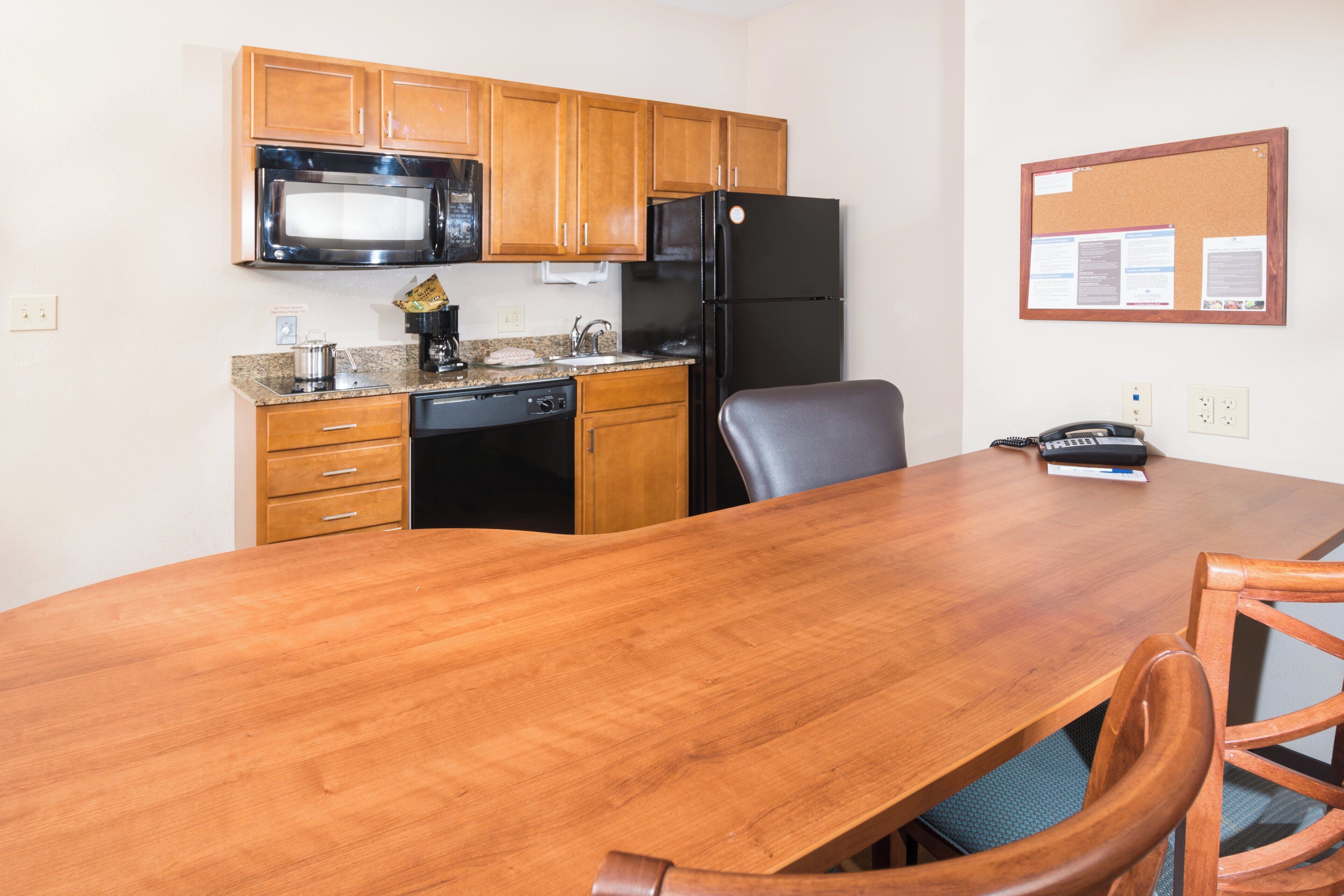 A compact kitchen with wooden cabinets, a black refrigerator, a microwave, and a coffee maker. There is a wooden dining table with chairs, and a corkboard with notices on the wall.