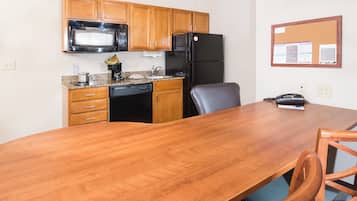 A compact kitchen with wooden cabinets, a black refrigerator, a microwave, and a coffee maker. There is a wooden dining table with chairs, and a corkboard with notices on the wall.