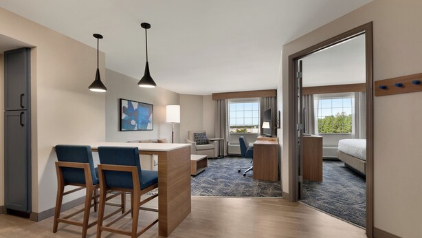 A modern hotel room with a kitchenette, a dining area with two bar stools, a desk with a chair, a large bed, and a view of the outdoors.