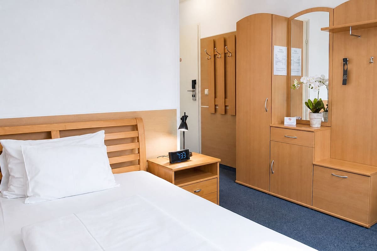 A hotel room with a bed, a bedside table with a digital clock, a nightstand, a wardrobe, and a mirror.