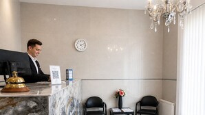 A hotel reception area with a marble counter, a receptionist, two black chairs, a clock, and a chandelier.